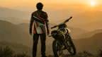 Person standing next to a motorcycle with a sunset and mountainous landscape, featuring the quote 'Less Traveled. More Lived.