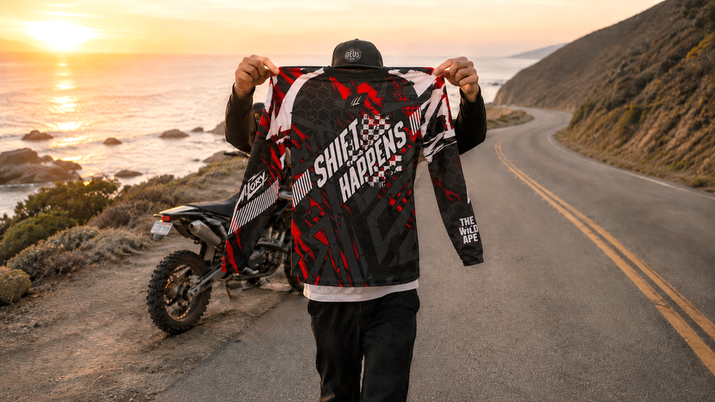 Person holding a jacket with 'Shift Happens' text on a scenic road with ocean view
